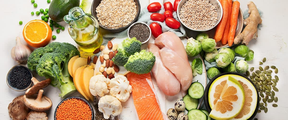 Verschiedene gesunde Lebensmittel wie Hähnchenbrust, Lachs, Brokkoli, Blumenkohl, Tomaten, Gurken, Karotten, Rosenkohl, Quinoa, Haferflocken, Chiasamen, Mandeln, Linsen, Wachteleier, Birne, Zitrone und eine Flasche Olivenöl