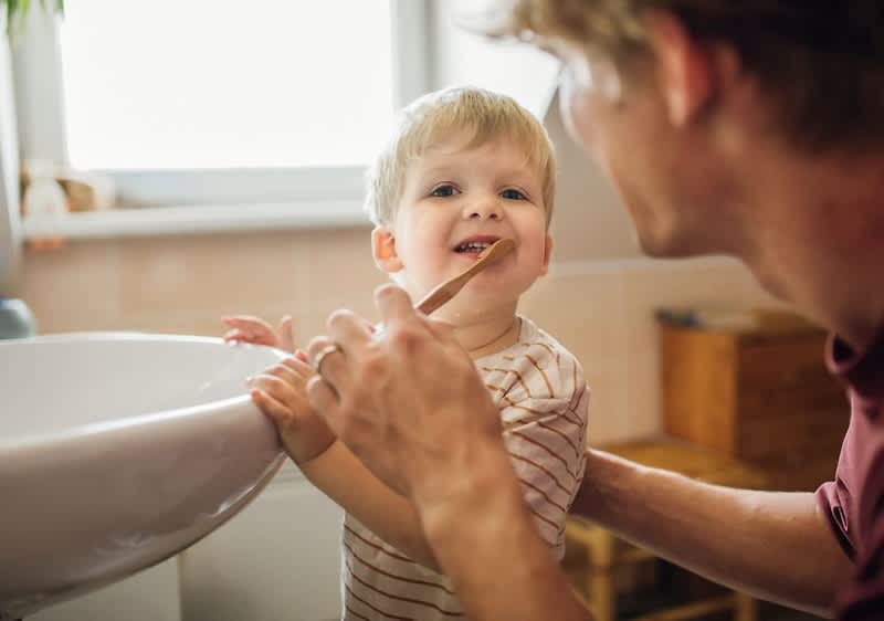Ein Kleinkind steht an einem weißen Waschbecken und wird von einem Erwachsenen unterstützt, der eine Hand am Rücken des Kindes hat