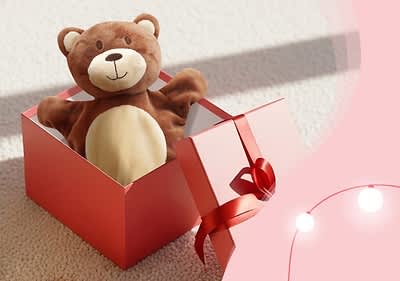 Ein brauner Plüschbär mit einem lächelnden Gesicht sitzt in einer geöffneten roten Geschenkbox mit roter Schleife auf einem hellen Teppich