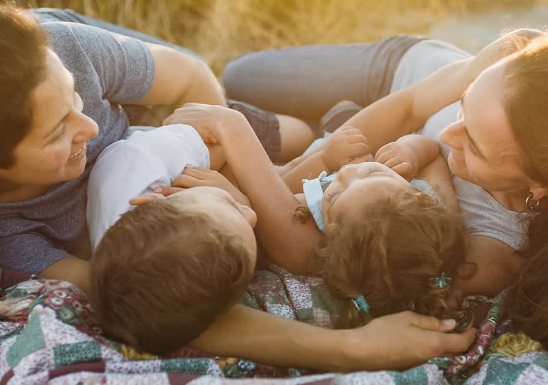 Eine Familie liegt entspannt auf einer bunten Decke im Freien, die Erwachsenen lächeln und schauen liebevoll auf die Kinder