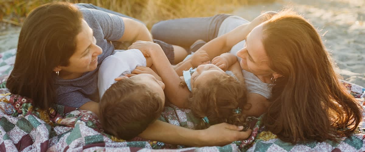 Eine Familie liegt entspannt auf einer bunten Decke im Freien, die Erwachsenen lächeln und schauen liebevoll auf die Kinder
