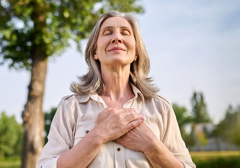 Grauhaarige Frau mit gefalteten Händen auf der Brust atmet tief ein