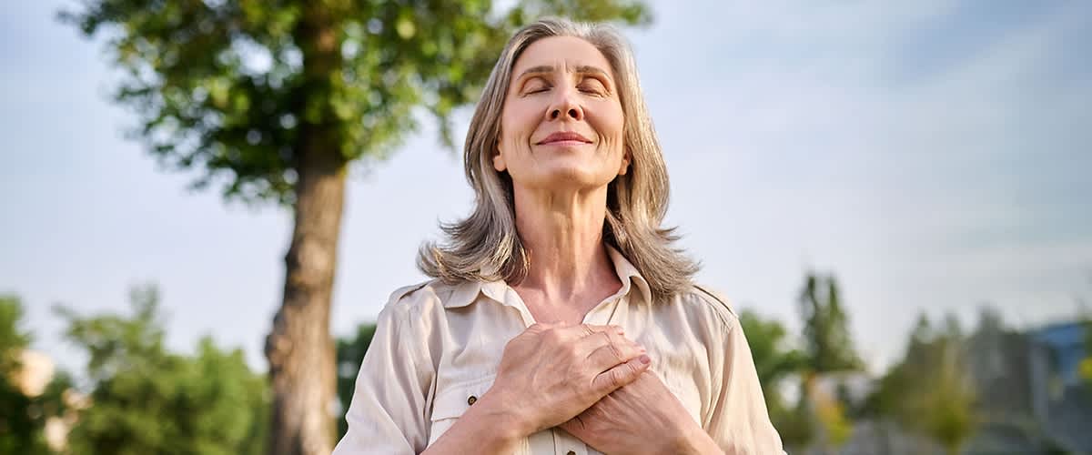 Grauhaarige Frau mit gefalteten Händen auf der Brust atmet tief ein