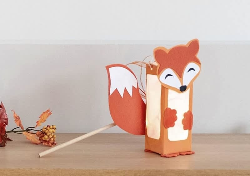 Eine Laterne in Form eines Fuchses mit orangefarbenem Körper und weißem Gesicht, Schwanz und Pfoten steht auf einem Holztisch