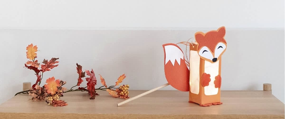 Eine Laterne in Form eines Fuchses mit orangefarbenem Körper und weißem Gesicht, Schwanz und Pfoten steht auf einem Holztisch