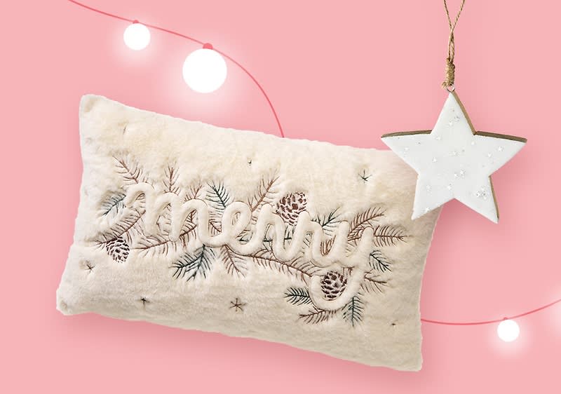 Ein cremefarbenes Kissen mit gesticktem Schriftzug „merry“ und Tannenzapfen-Motiv, daneben hängt ein weißer Stern aus Holz an einer Schnur