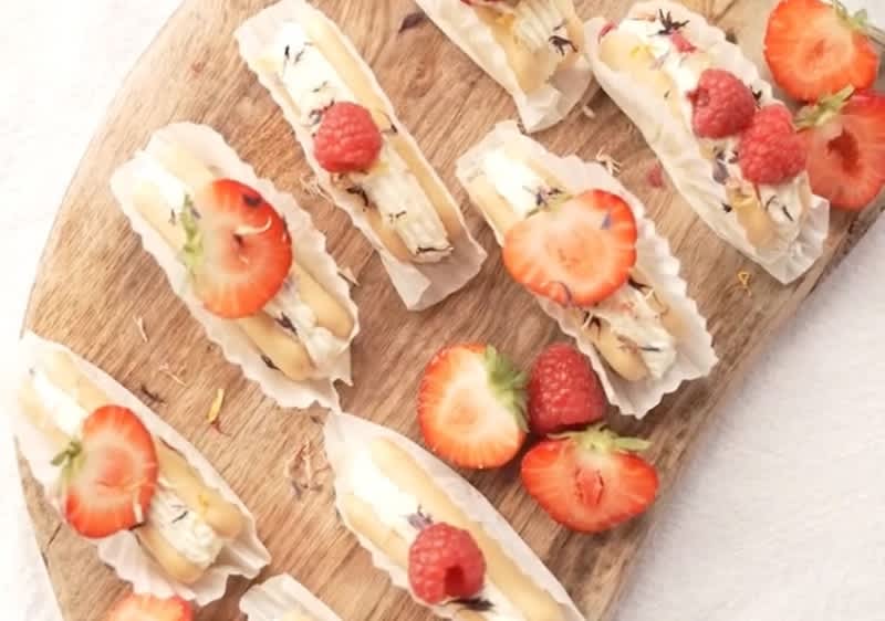 Mehrere längliche Gebäckstücke mit Sahnefüllung, garniert mit frischen Himbeeren und Erdbeerscheiben, liegen auf einem runden Holzbrett