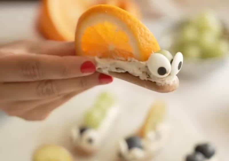Eine Hand mit rotem Nagellack hält einen Snack in Form einer Schnecke, bestehend aus einer Orangenscheibe als Schneckenhaus und zwei Augen aus weißer Creme mit schwarzen Punkten auf einem Keks