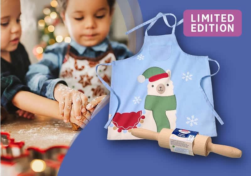 Zwei Kinder backen Kekse, daneben eine blaue Kinderschürze mit einem Weihnachtsbärenmotiv und ein Nudelholz mit der Aufschrift „LIMITED EDITION“
