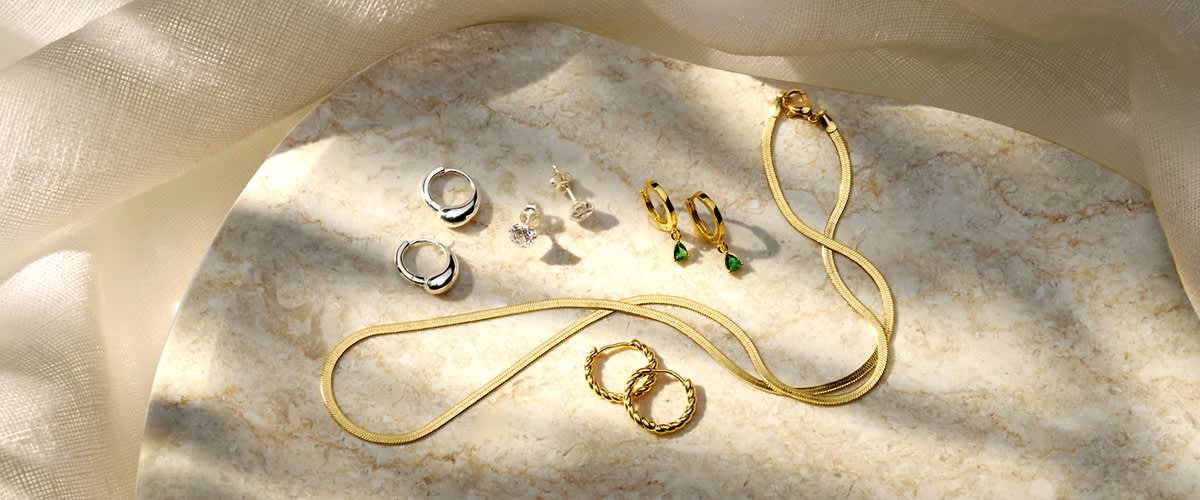 Goldene Kette, zwei goldene Ringe mit Zierkante, zwei kleine goldene Creolen mit grünen Anhängern, zwei silberne Creolen und zwei Paar Steckerohrringe (kleine runde Steine) auf hellem Marmoruntergrund