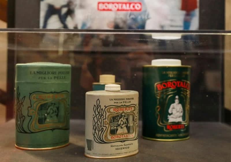 Drei Borotalco-Puderbehälter in verschiedenen Größen und Farben, vorne ein kleiner weißer Behälter mit der Aufschrift „La migliore polvere per la pelle“, dahinter ein grüner und ein dunkelgrüner Behälter mit ähnlichen Aufschriften, im Hintergrund ein verschwommenes Borotalco-Logo