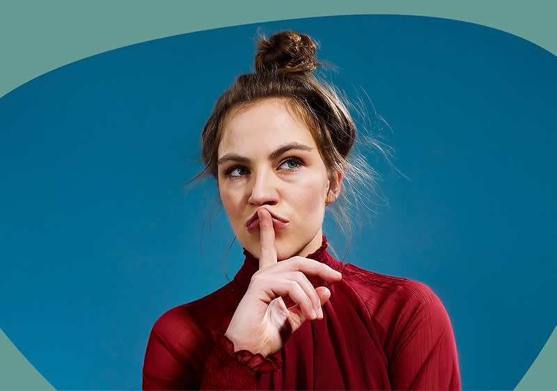 Eine Person in einem roten Oberteil hält einen Finger vor den Mund, der Hintergrund ist blau