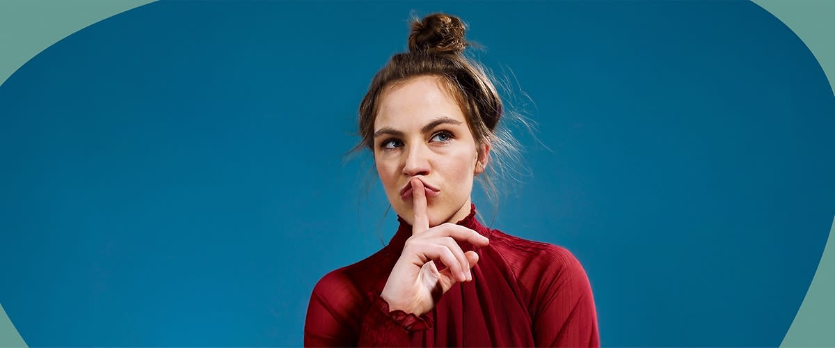 Eine Person in einem roten Oberteil hält einen Finger vor den Mund, der Hintergrund ist blau