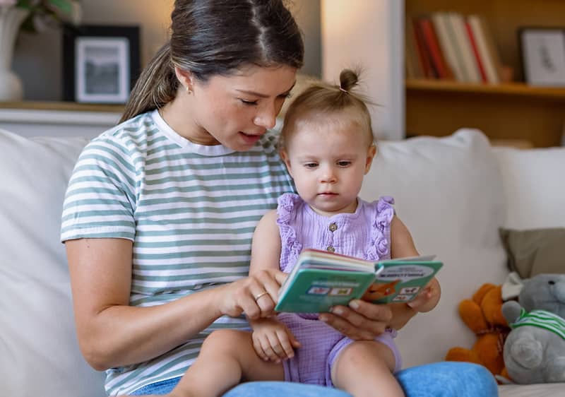 Eine Frau in einem gestreiften T-Shirt sitzt auf einem Sofa und liest einem Kleinkind in einem lila Strampler ein Buch vor, neben ihnen liegt ein graues Stofftier