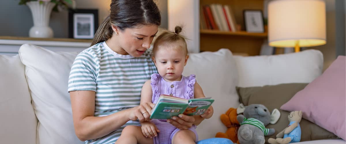 Eine Frau in einem gestreiften T-Shirt sitzt auf einem Sofa und liest einem Kleinkind in einem lila Strampler ein Buch vor, neben ihnen liegt ein graues Stofftier