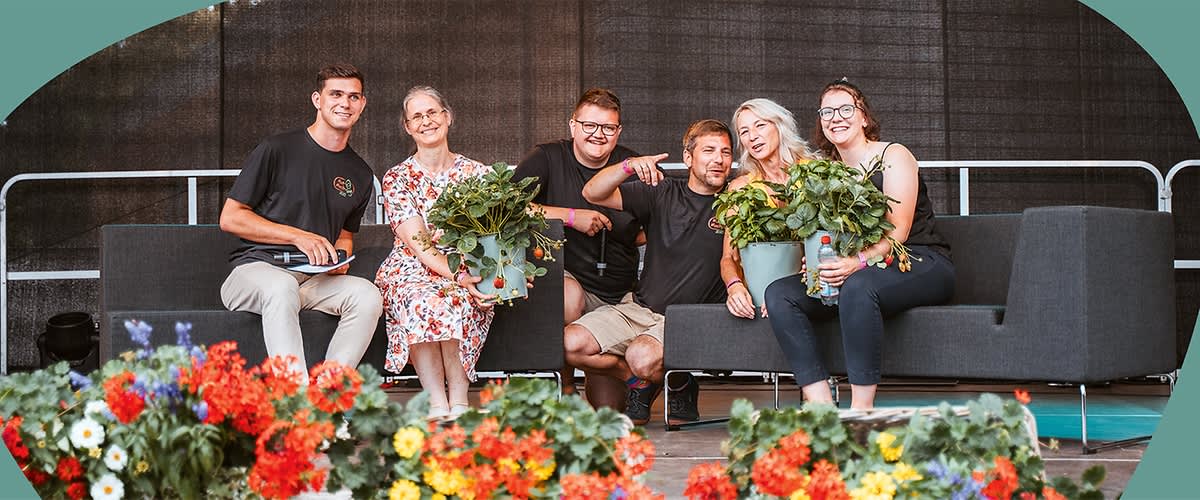 Eine Gruppe von fünf Personen sitzt auf einer Bühne, drei von ihnen halten grüne Pflanzen in Töpfen, im Vordergrund sind bunte Blumen zu sehen