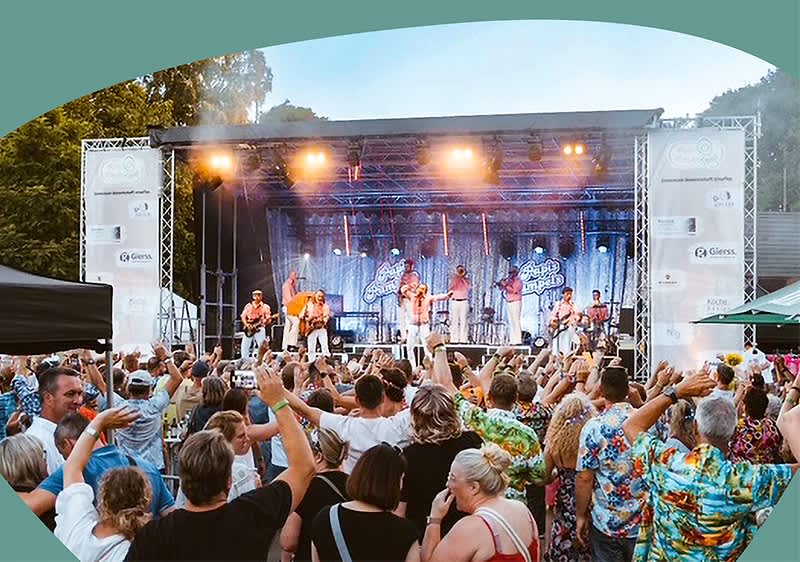 Eine Menschenmenge jubelt vor einer Bühne, auf der eine Band mit mehreren Musikern spielt, umgeben von bunten Lichtern und transparenten Vorhängen