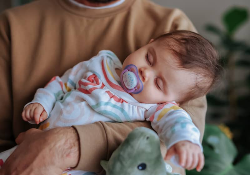 Ein Baby in einem bunten Strampler mit Regenbogenmuster liegt in den Armen eines Erwachsenen, der einen braunen Pullover trägt, neben dem Baby ein grünes Stofftier