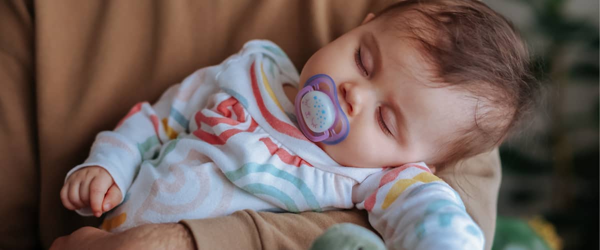Ein Baby in einem bunten Strampler mit Regenbogenmuster liegt in den Armen eines Erwachsenen, der einen braunen Pullover trägt, neben dem Baby ein grünes Stofftier