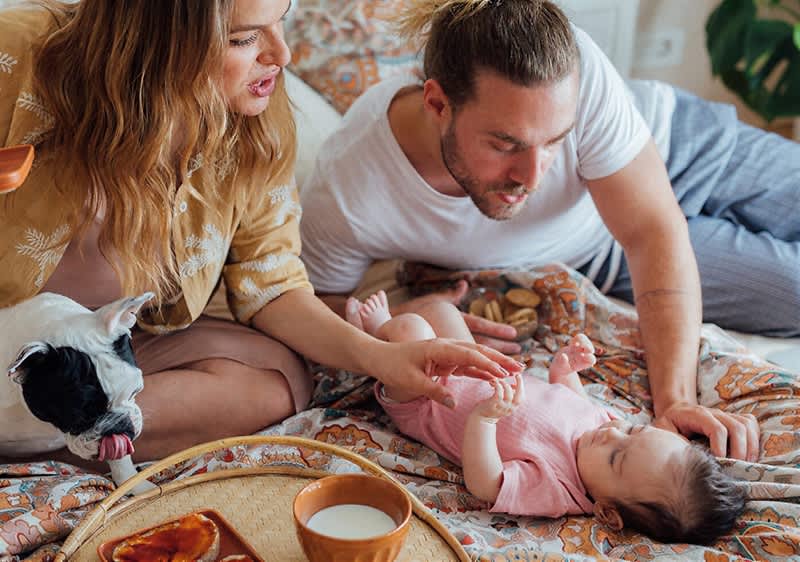 Ein Baby in einem rosa Strampler liegt auf einem Bett, während eine Frau mit einem Hund und ein Mann daneben sitzen. Ein Frühstückstablett mit einer Tasse und einem Teller ist im Vordergrund sichtbar.