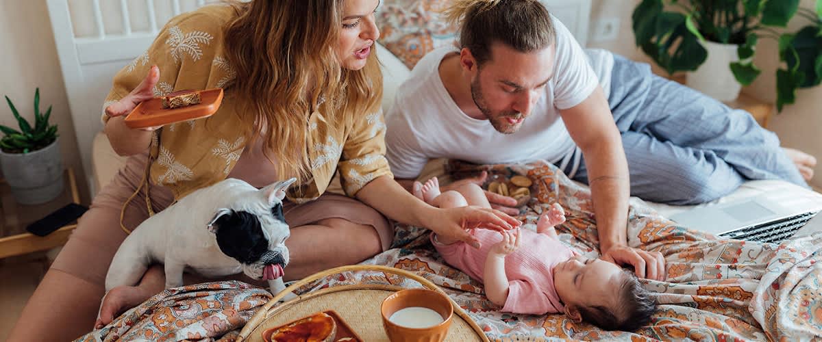 Ein Baby in einem rosa Strampler liegt auf einem Bett, während eine Frau mit einem Hund und ein Mann daneben sitzen. Ein Frühstückstablett mit einer Tasse und einem Teller ist im Vordergrund sichtbar.