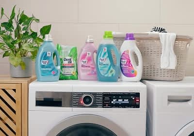 Fünf Flaschen Waschmittel der Marke Denkmit in verschiedenen Farben stehen auf einer Waschmaschine, daneben ein Wäschekorb mit weißen Handtüchern und eine grüne Pflanze in einem Topf