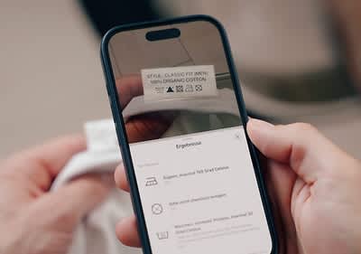 Ein Smartphone scannt ein Pflegeetikett mit der Aufschrift „Style Classic Fit Men 100% Organic Cotton“ und zeigt die Waschanleitung auf dem Bildschirm an, darunter „Ergebnisse“, „Bügeln, maximal 150 Grad Celsius“ und „Nicht chemisch reinigen“
