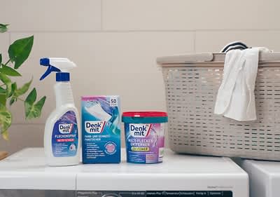 Eine weiße Sprühflasche mit der Aufschrift „Denk mit Fleckenspray“, daneben eine blaue Verpackung mit der Aufschrift „Denk mit Farb- und Schmutz-Fangtücher“ und ein roter Behälter mit der Aufschrift „Denk mit Kraft-Fleckentferner“ stehen auf einer Waschmaschine, daneben ein Wäschekorb mit einem weißen Hemd