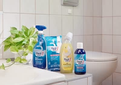 Vier Reinigungsprodukte der Marke Denk mit stehen auf einem Badezimmerregal, darunter Badreiniger, Reinigungstücher, Reinigungsgel und Allzweckreiniger, daneben eine grüne Pflanze