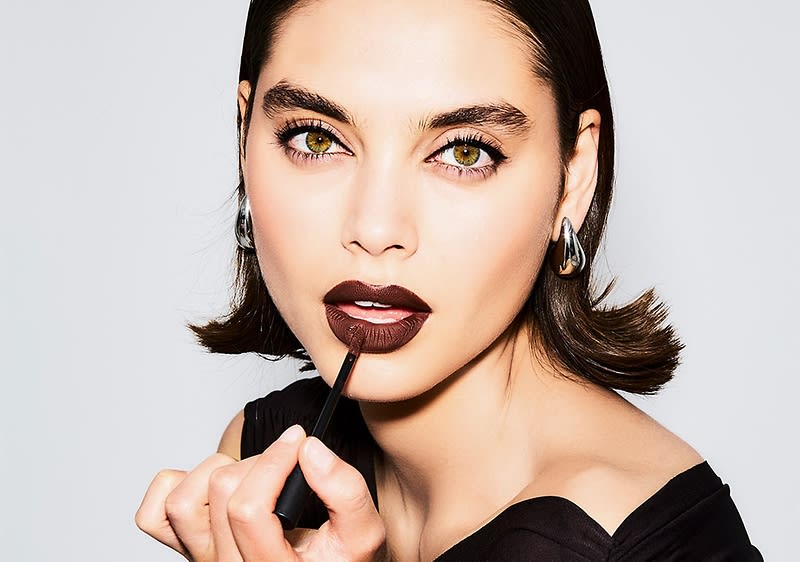 Eine Person mit kurzen braunen Haaren trägt silberne Ohrringe und hält einen Lippenstift in der Hand