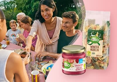 Eine Gruppe von Menschen sitzt an einem Tisch und genießt eine Mahlzeit. Im Vordergrund sind ein Glas Bio-Sirup mit der Aufschrift „Bio Sirup Holunderblüte“ und eine Packung Bio-Winternudeln mit bunten Nudeln zu sehen.