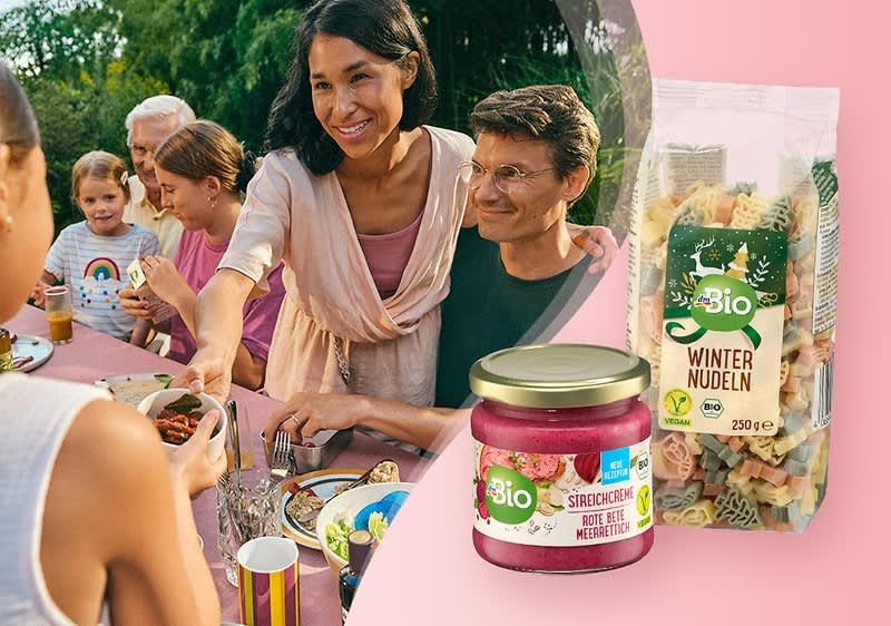 Eine Gruppe von Menschen sitzt an einem Tisch und genießt eine Mahlzeit. Im Vordergrund sind ein Glas Bio-Sirup mit der Aufschrift „Bio Sirup Holunderblüte“ und eine Packung Bio-Winternudeln mit bunten Nudeln zu sehen.