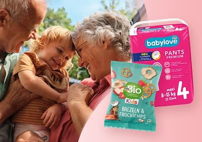 Eine ältere Person hält ein Kleinkind im Arm. Daneben sind eine Packung Babylove Pants Premium und eine Tüte Bio Kids Brezeln & Fruchtchips zu sehen.