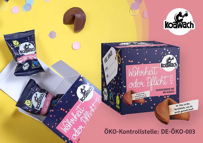 Eine dunkelblaue Verpackung mit der Aufschrift „Wahrheit oder Pflicht? Schokokeks mit Bio-Kakao“ und dem Koawach-Logo. Daneben liegen zwei geöffnete Keksverpackungen mit dem gleichen Logo. Ein Glückskeks ist geöffnet, und ein Zettel mit der Aufschrift „Wie wäre es mit einem Schokokeks?“ ist sichtbar. ÖKO-Kontrollstelle: DE-ÖKO-003
