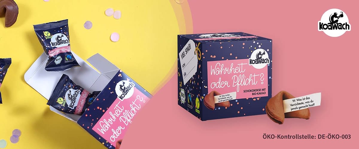 Eine dunkelblaue Verpackung mit der Aufschrift „Wahrheit oder Pflicht? Schokokeks mit Bio-Kakao“ und dem Koawach-Logo. Daneben liegen zwei geöffnete Keksverpackungen mit dem gleichen Logo. Ein Glückskeks ist geöffnet, und ein Zettel mit der Aufschrift „Wie wäre es mit einem Schokokeks?“ ist sichtbar. ÖKO-Kontrollstelle: DE-ÖKO-003