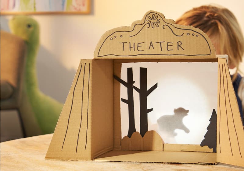 Ein Kind spielt mit einem selbstgebauten Pappkarton-Theater, das den Schriftzug „THEATER“ trägt und eine Schattenfigur eines Bären vor zwei Bäumen zeigt