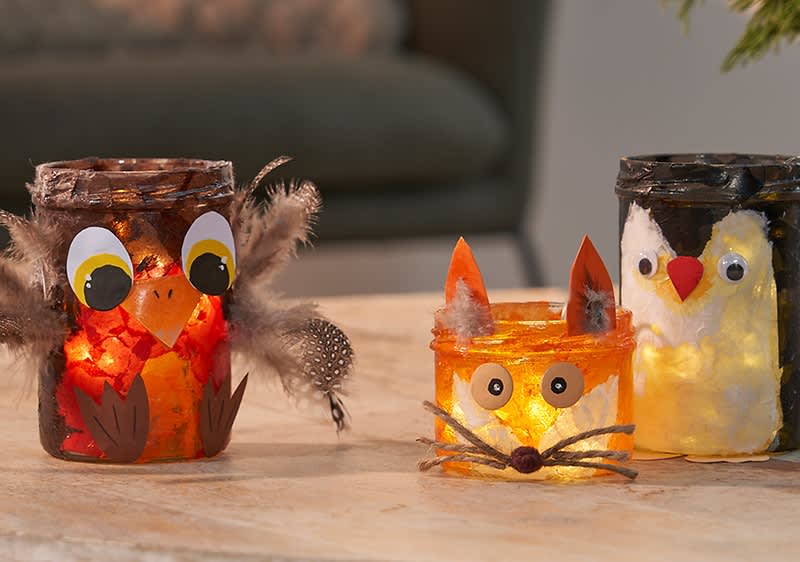 Drei dekorierte Gläser mit Teelichtern, links ein Glas mit Eulenmotiv und Federn, in der Mitte ein Glas mit Fuchsmotiv und Schnurrhaaren, rechts ein Glas mit Pinguinmotiv