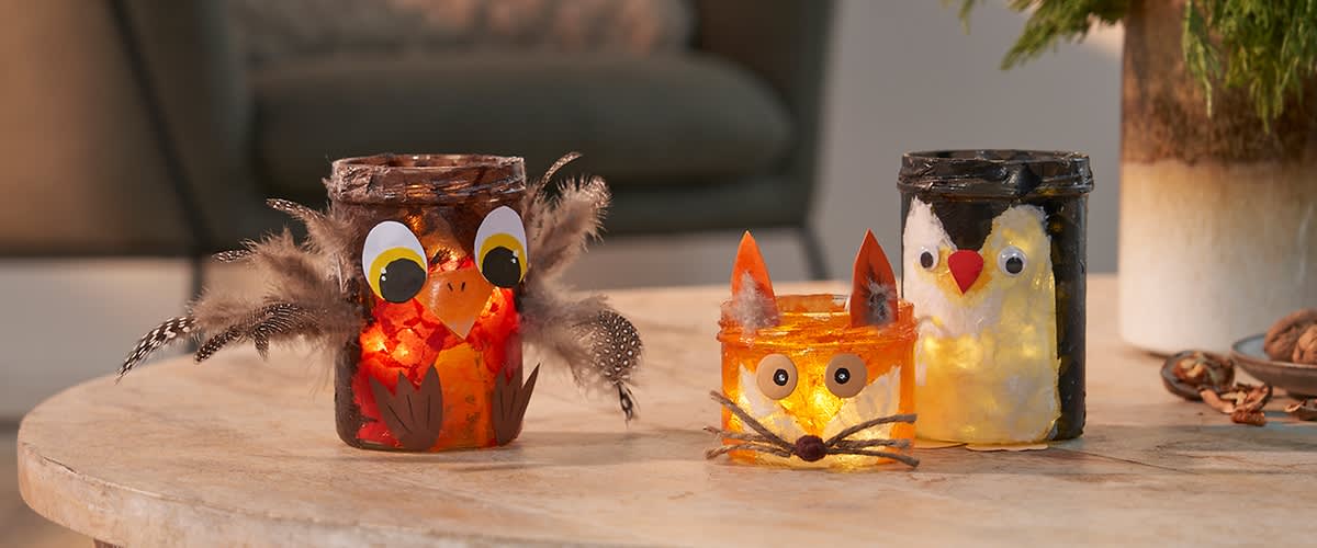 Drei dekorierte Gläser mit Teelichtern, links ein Glas mit Eulenmotiv und Federn, in der Mitte ein Glas mit Fuchsmotiv und Schnurrhaaren, rechts ein Glas mit Pinguinmotiv