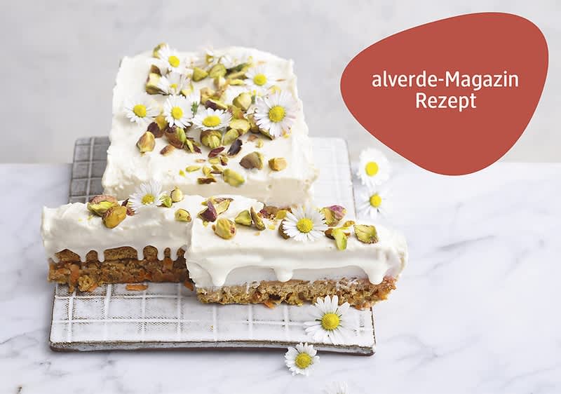 Zwei eckige Karottenkuchenstücke mit weißem Topping aus Joghurt, oben mit gehackten Pistazien und kleinen Gänseblümchen dekoriert, auf einem weißen Gitterrost und hellem Marmoruntergrund; oben rechts ein roter, organisch geformter Schriftkreis mit weißer Aufschrift „alverde-Magazin Rezept“