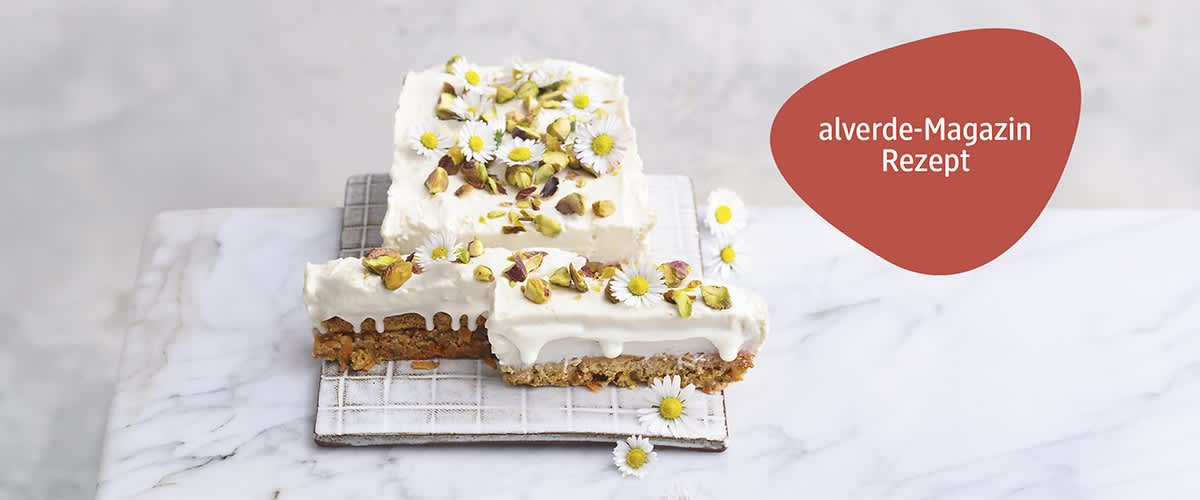 Zwei eckige Karottenkuchenstücke mit weißem Topping aus Joghurt, oben mit gehackten Pistazien und kleinen Gänseblümchen dekoriert, auf einem weißen Gitterrost und hellem Marmoruntergrund; oben rechts ein roter, organisch geformter Schriftkreis mit weißer Aufschrift „alverde-Magazin Rezept“