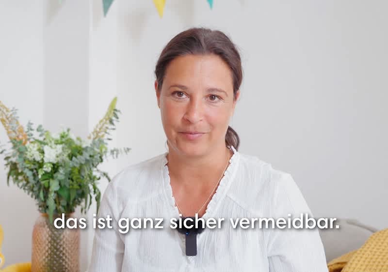 Eine Frau in einem weißen Oberteil sitzt vor einem Hintergrund mit Pflanzen und bunten Wimpeln, der Text „das ist ganz sicher vermeidbar“ ist eingeblendet.