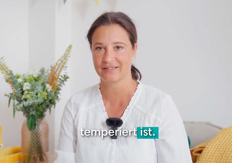 Eine Person in einem weißen Hemd trägt ein Mikrofon, daneben ein Blumenstrauß in einer Vase, der Text „temperiert ist“ ist eingeblendet