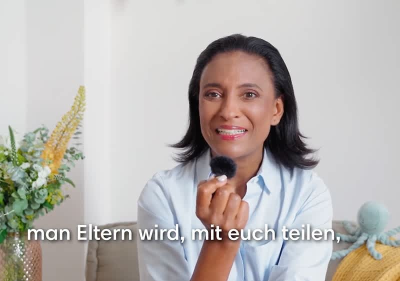 Dr. Karella Easwaran hält ein Mikrofon, im Hintergrund sind Pflanzen zu sehen, der Text lautet: „man Eltern wird, mit euch teilen,“