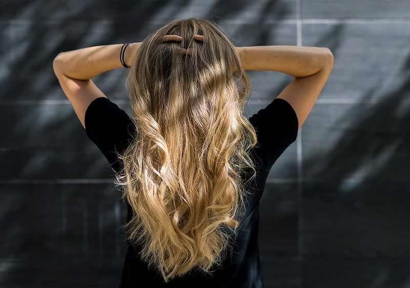 Eine Person mit langen, welligen Haaren steht mit dem Rücken zur Kamera und hält ihre Hände am Kopf