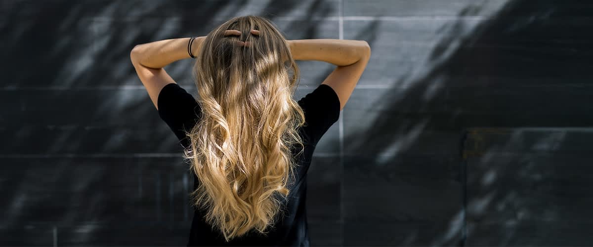 Eine Person mit langen, welligen Haaren steht mit dem Rücken zur Kamera und hält ihre Hände am Kopf