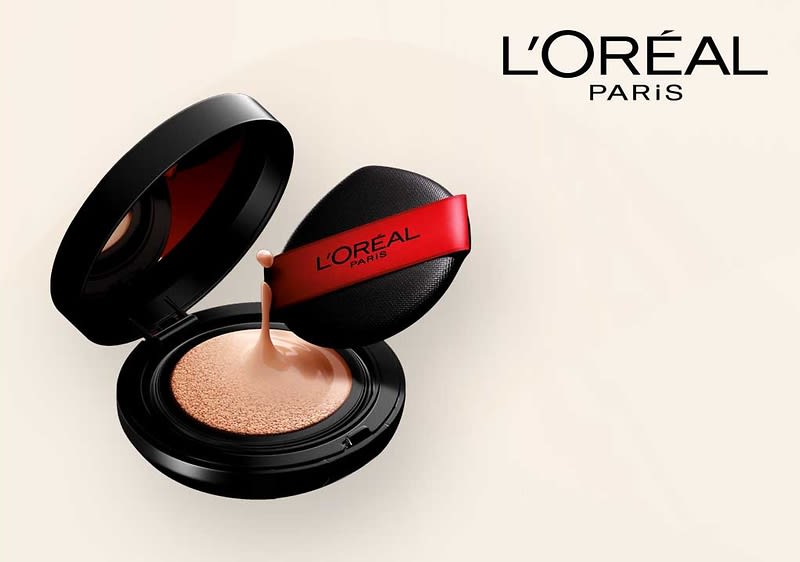 Eine schwarze Puderdose mit offenem Deckel zeigt eine beige Foundation, daneben ein schwarzer Applikator mit rotem Band und der Aufschrift „L'Oréal Paris“, oben rechts das Logo von L'Oréal Paris