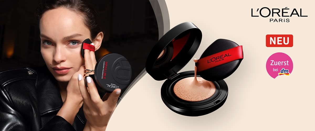 Eine schwarze Puderdose mit offenem Deckel zeigt eine beige Foundation, daneben ein schwarzer Applikator mit rotem Band und der Aufschrift „L'Oréal Paris“, darüber steht „NEU“ in einem roten Kasten, daneben „Zuerst bei dm“ in einem rosa Kreis, oben rechts das Logo von L'Oréal Paris