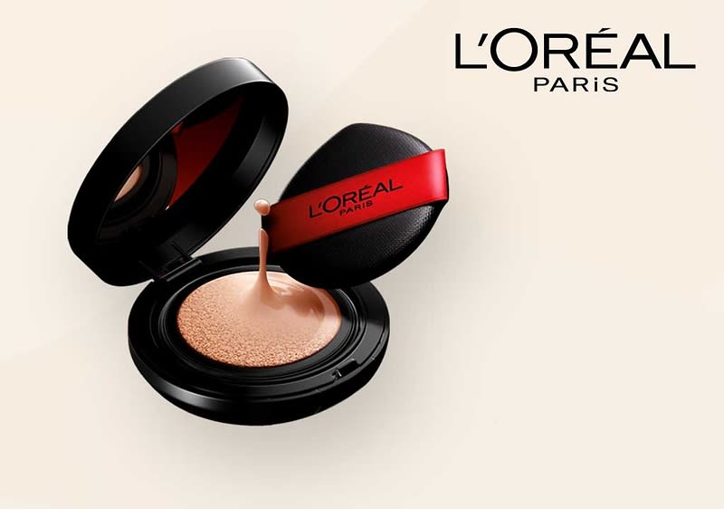 Eine schwarze Puderdose mit offenem Deckel zeigt eine beige Foundation, daneben ein schwarzer Applikator mit rotem Band und der Aufschrift „L'Oréal Paris“, oben rechts das Logo von L'Oréal Paris