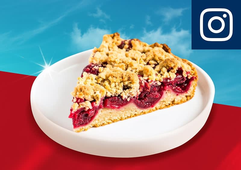 Ein Stück Kirsch-Streuselkuchen auf einem weißen Teller vor einem Hintergrund aus blauem Himmel und rotem Boden, oben rechts das Instagram-Logo