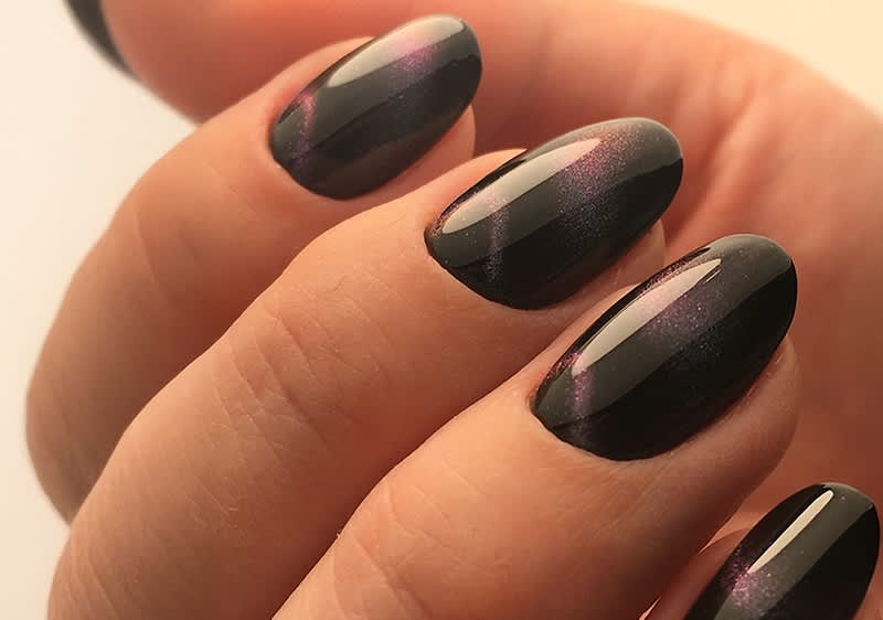 Nahaufnahme einer Hand mit ovalen, dunkel lackierten Fingernägeln, die einen schimmernden violetten Akzent haben